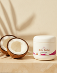 Coconut Cloud Whipped Body Butter on travertine beside two coconut halves under palm-leaf shadows