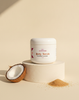 Coconut Cloud Whipped Sugar Scrub on a cream pedestal with a halved coconut and golden sugar