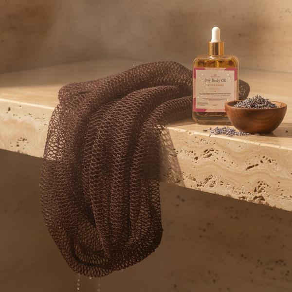A cocoa-brown soft African exfoliating net draped over warm travertine beside a bottle of dry body oil and a bowl of lavender buds