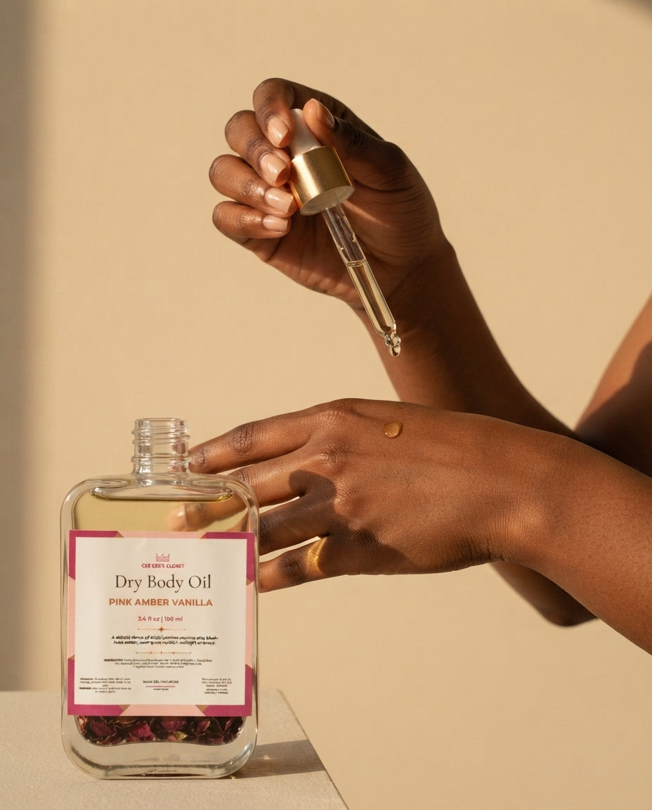A hand holding a gold dropper above a bottle of Pink Amber Vanilla Dry Body Oil with hibiscus petals inside, a drop of oil on the back of the other hand