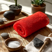 Red african exfoliating net, stones, and a wooden tool on a bamboo mat with plants in the background for peaceful gentle exfoliation towards softer and smoother skin