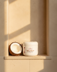 Coconut Cloud Whipped Sugar Scrub in a sunlit shower niche beside a halved coconut, with water droplets on the jar