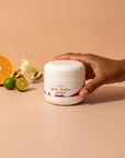 Hand resting on a Galaxy Glow Whipped Body Butter jar with orange, limequats and freesia