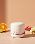 Galaxy Glow Whipped Body Butter jar on a peach backdrop with halved orange, limequats, rhubarb and freesia