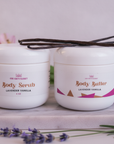 Soft Skin Set in Lavender Vanilla – Body Scrub and Body Butter topped with vanilla beans among pink lilies